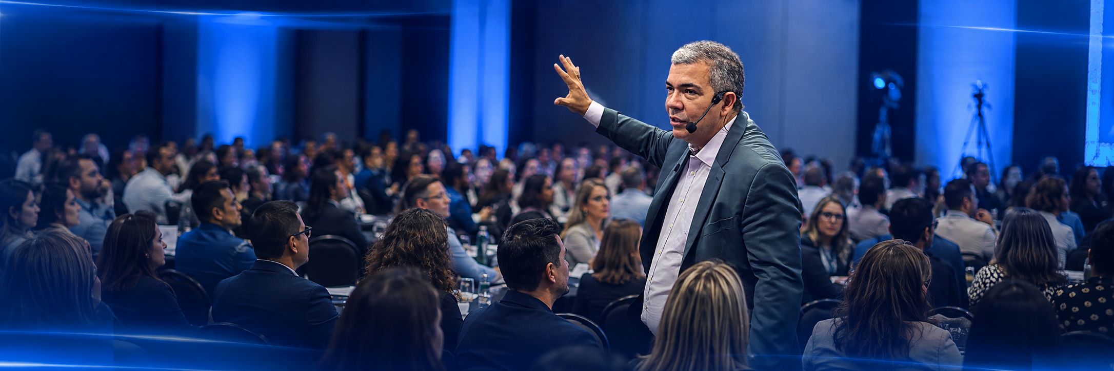 Newton Vieira em pé no meio da plateia, de microfone headset, falando com a sala cheia